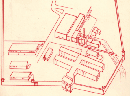 Isometric red line drawing of a walled industrial or institutional complex. The compound contains several rectangular buildings, including long barracks-like structures, smaller auxiliary buildings, and what appears to be a central administrative or entry building at the front. The layout includes internal roads and a separate area with large buildings, possibly for manufacturing or processing, connected by a railway line. The perimeter is enclosed by walls with guard towers at each corner.